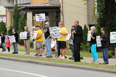 A group of parishioners stand along the side of the road holding signs.